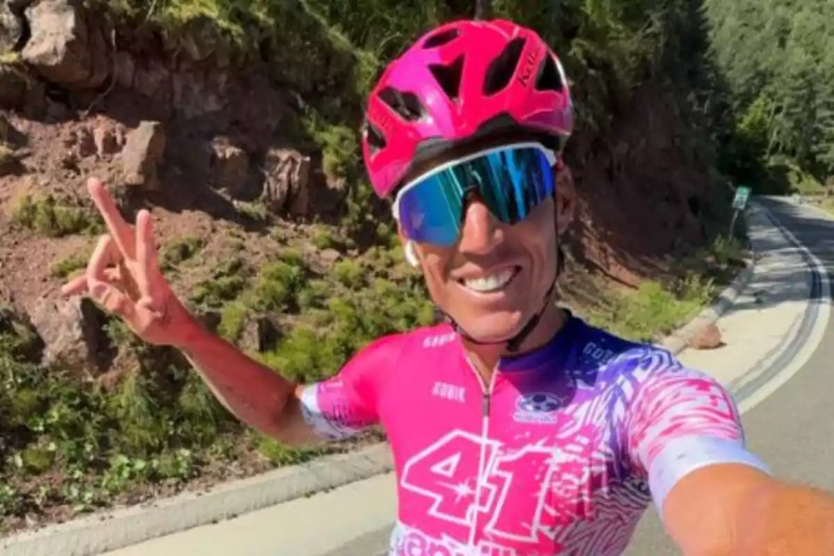 Ciclista sonriente con casco y gafas de sol en una carretera rodeada de naturaleza.