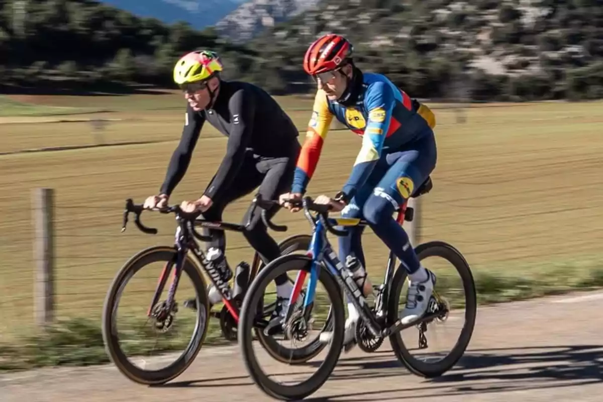 Dos ciclistas con cascos de colores brillantes pedalean juntos en una carretera rural rodeada de campos y montañas.