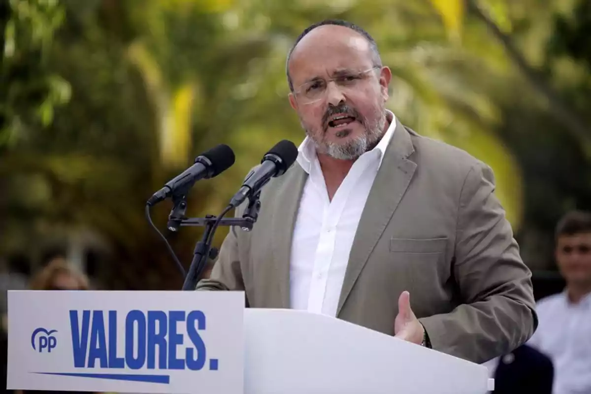 Hombre con barba y gafas hablando en un podio con la palabra VALORES y dos micrófonos al aire libre con fondo de vegetación