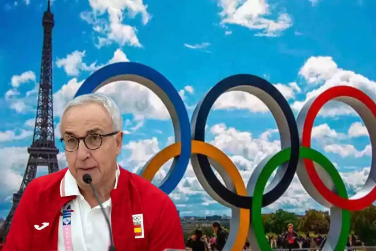 Un hombre con gafas y chaqueta roja habla frente a un micrófono con la Torre Eiffel y los anillos olímpicos de fondo.