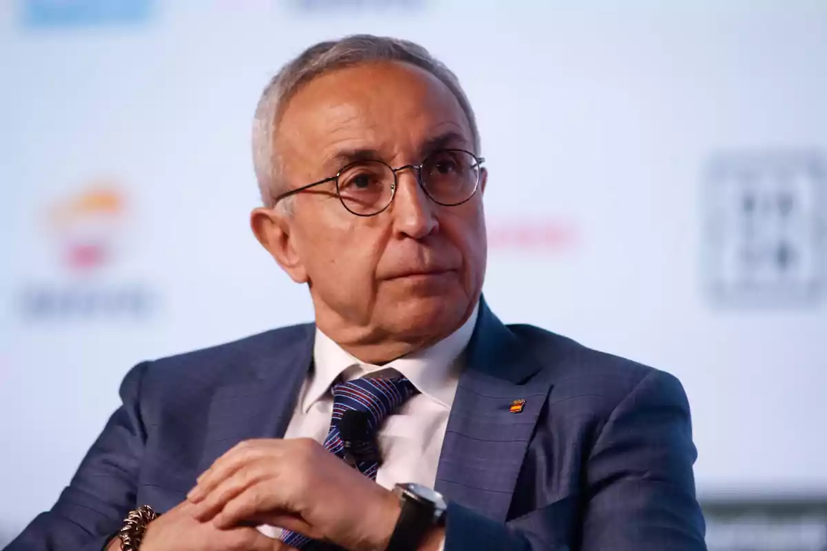 Un hombre mayor con gafas y traje azul claro, sentado y mirando hacia un lado en un evento.