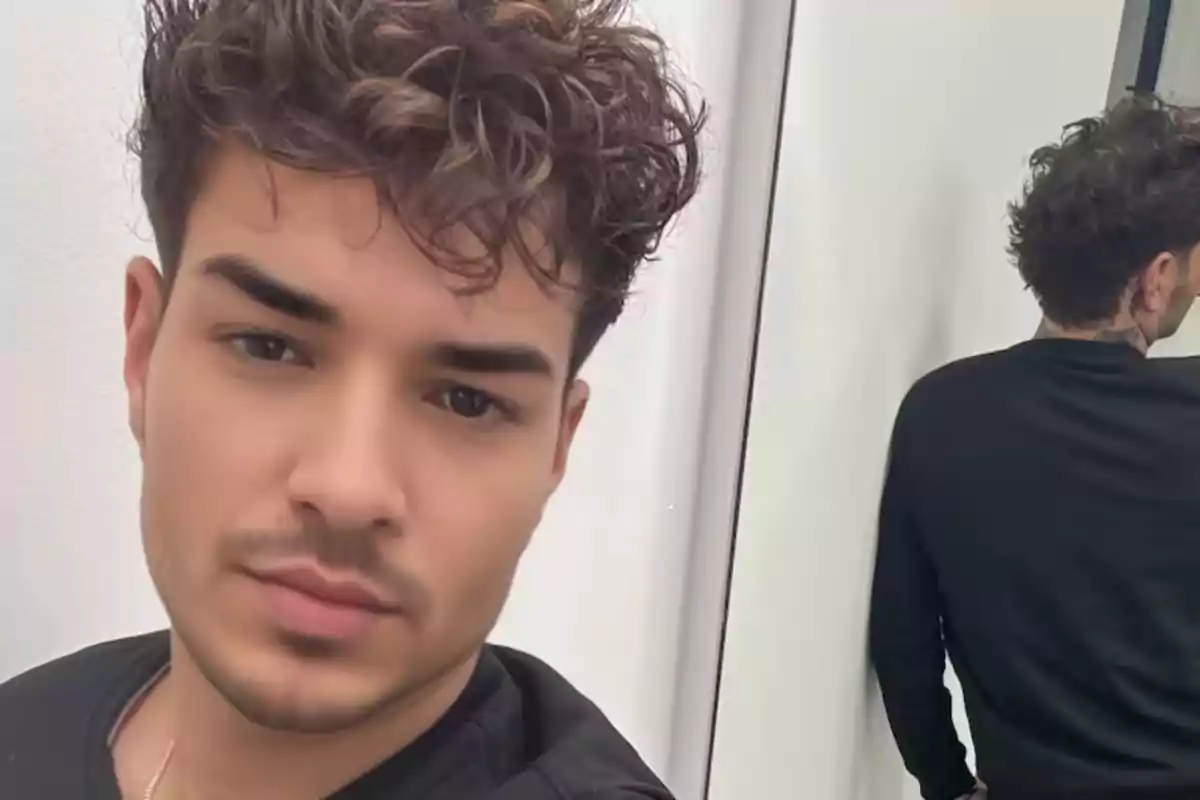 Un hombre con cabello rizado y barba ligera, vestido con una camiseta negra, se toma una selfie frente a un espejo.