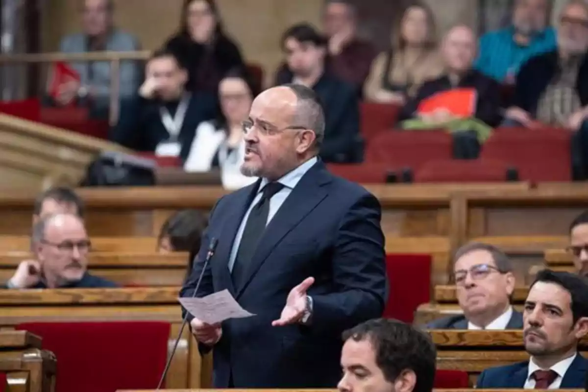 Hombre de traje hablando en un parlamento mientras sostiene unos papeles, rodeado de otras personas sentadas en bancas rojas Hombre de traje hablando en un parlamento mientras sostiene unos papeles, rodeado de otras personas sentadas en bancas rojas