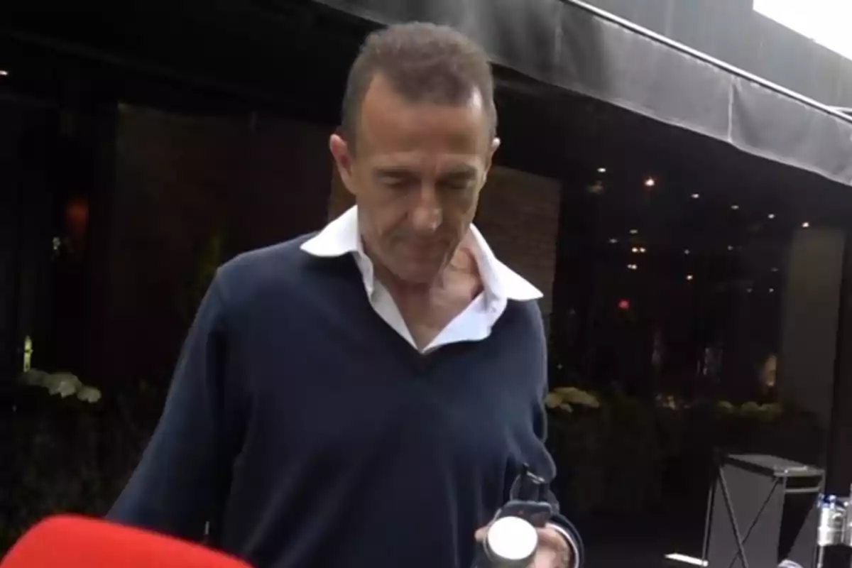 Alessandro Lequio con camisa blanca y suéter oscuro sostiene un objeto mientras camina frente a un edificio con luces. Alessandro Lequio con camisa blanca y suéter oscuro sostiene un objeto mientras camina frente a un edificio con luces.