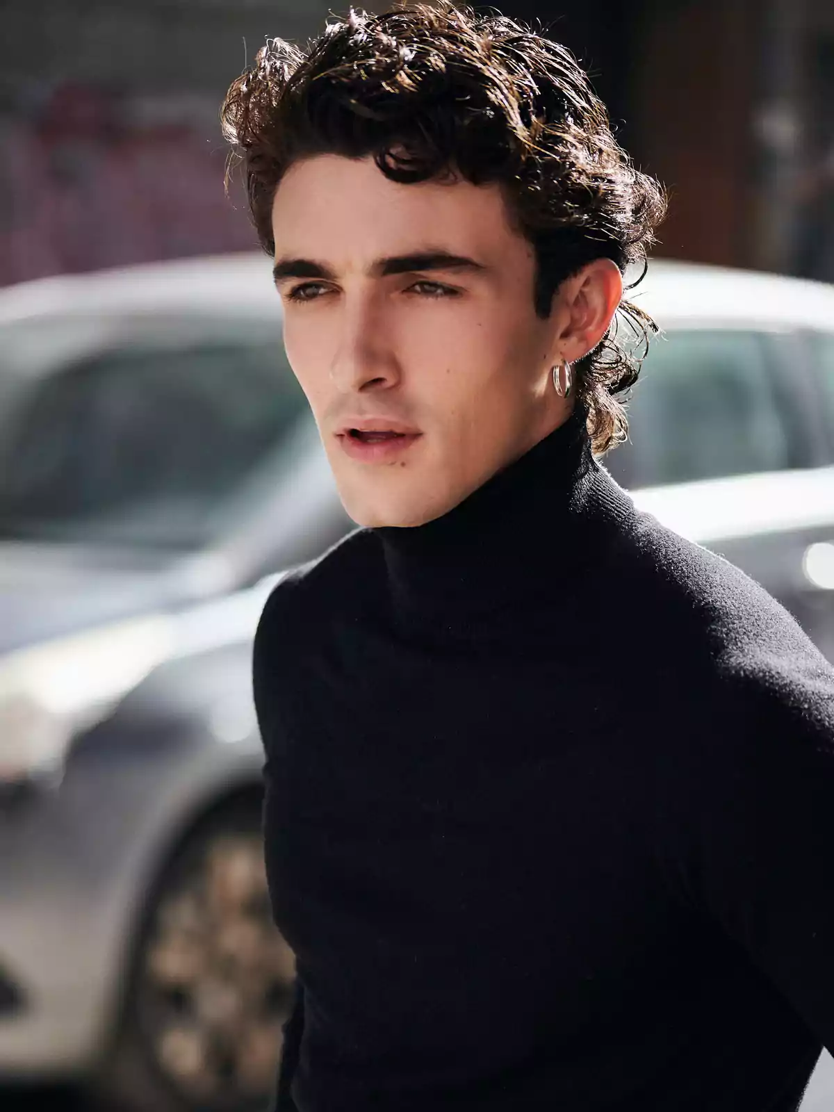 Un hombre con cabello rizado y oscuro, vistiendo un suéter negro de cuello alto, está de pie al aire libre con un coche desenfocado en el fondo.