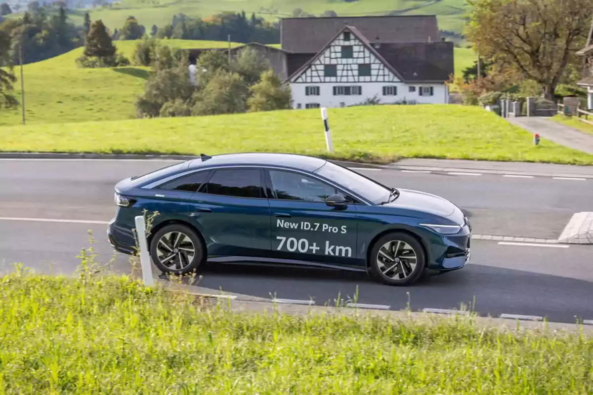 Un coche eléctrico Volkswagen ID.7 Pro S azul oscuro circula por una carretera rural con un paisaje verde y una casa de campo al fondo.