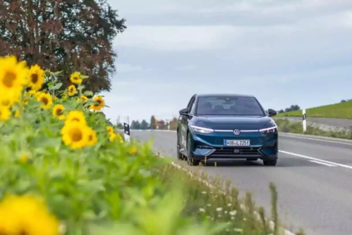 Un coche Volkswagen ID.7 Pro S azul circula por una carretera junto a un campo de girasoles bajo un cielo nublado.