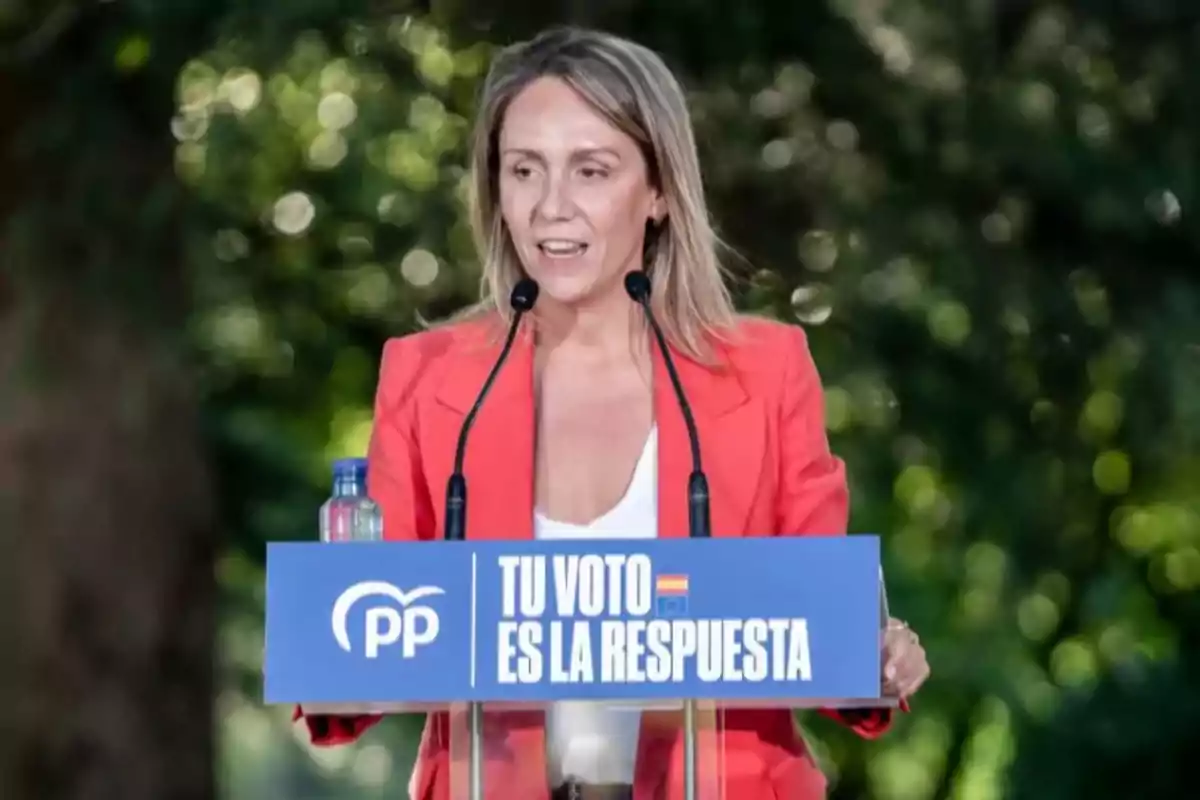 Mujer hablando en un mitin político al aire libre con un atril azul que tiene el logo del PP y el mensaje Tu voto es la respuesta