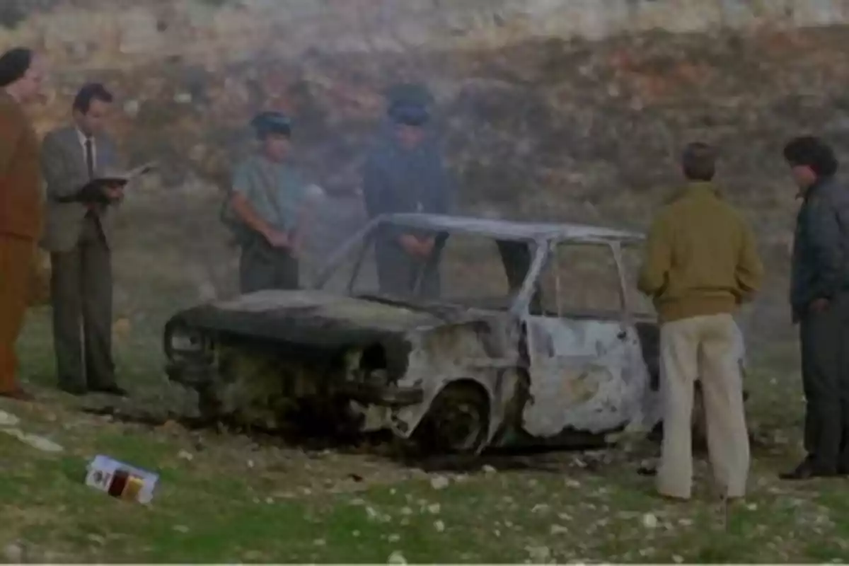 Un grupo de personas, incluyendo a dos oficiales, inspecciona un coche quemado en un entorno al aire libre.