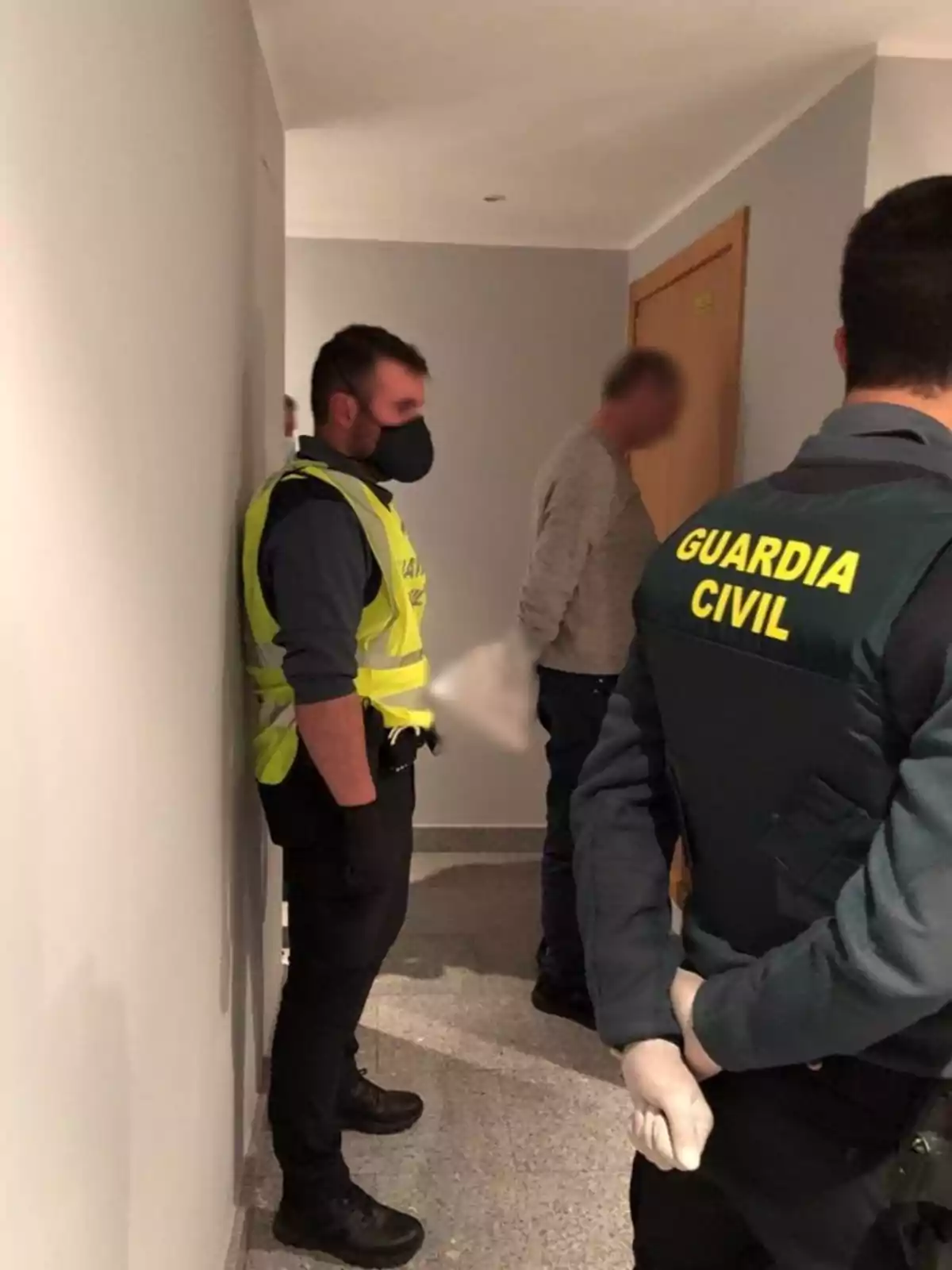 Dos agentes de la Guardia Civil con chalecos y mascarillas están de pie en un pasillo junto a una persona con el rostro difuminado.