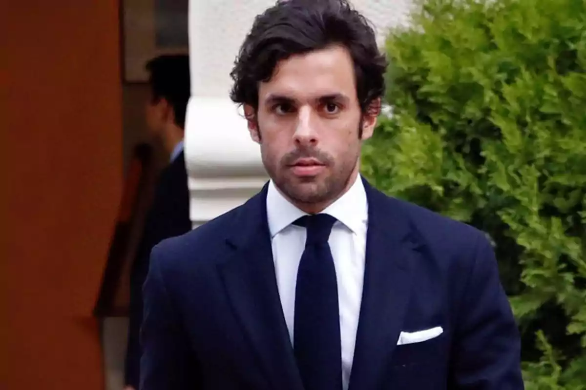 Un hombre con traje oscuro y corbata, de pie frente a un fondo con vegetación.