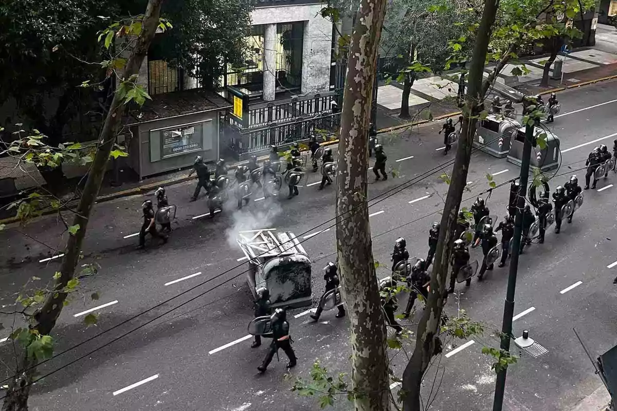 Un grupo de policías con equipo antidisturbios marcha por una calle junto a un contenedor de basura volcado y humeante, rodeados de árboles y edificios.