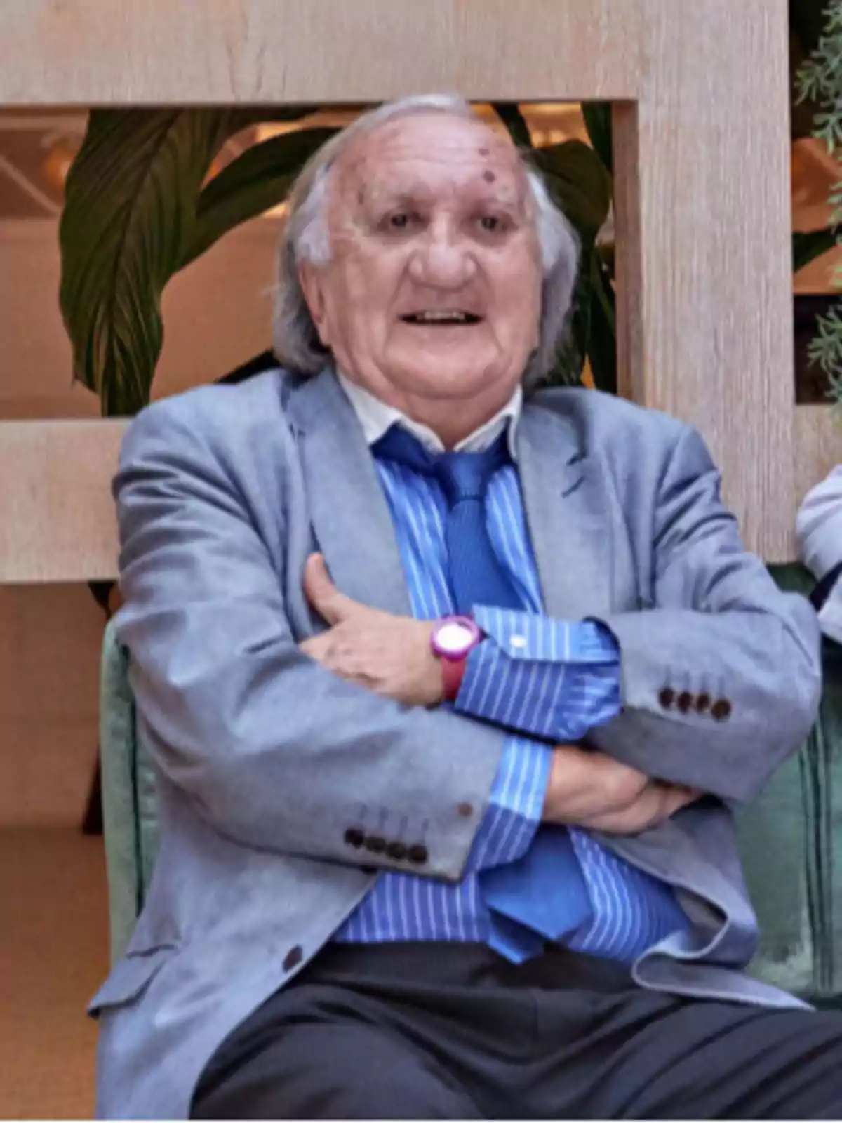 Un hombre mayor con cabello canoso y una chaqueta gris, camisa azul a rayas y corbata azul, está sentado con los brazos cruzados y sonriendo.