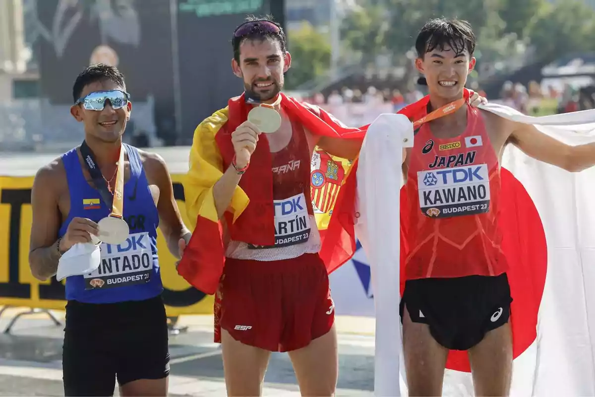 Tres atletas posan con sus medallas después de una competencia, uno de ellos envuelto en la bandera de España, otro con la bandera de Japón y el tercero con la bandera de Ecuador.