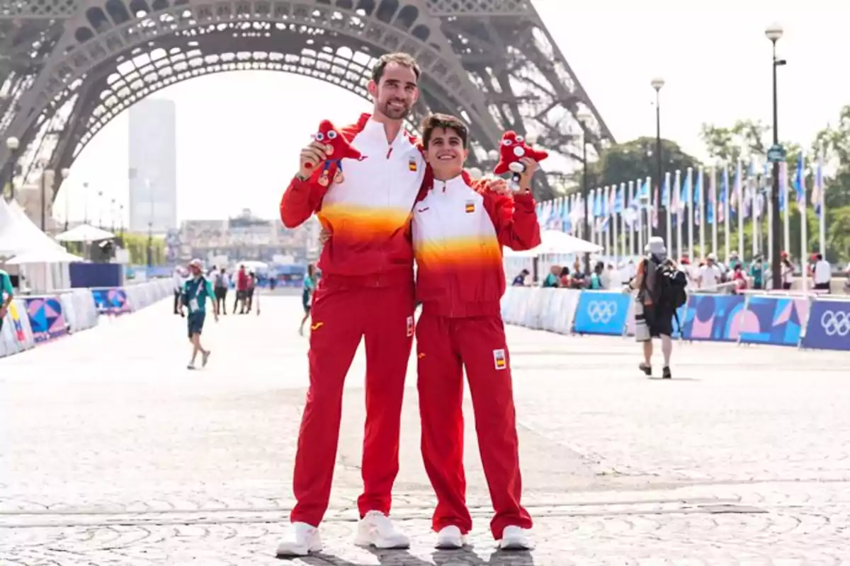 Dos personas con uniformes deportivos rojos y blancos posan frente a la Torre Eiffel sosteniendo peluches rojos.