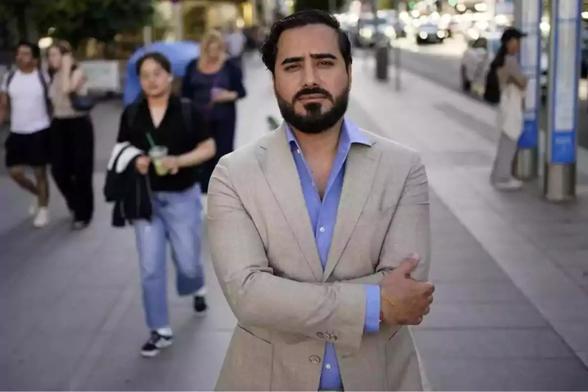 Hombre con barba y traje claro de pie en una calle concurrida con personas caminando en el fondo.