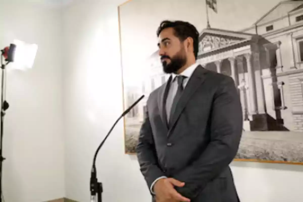 Hombre de traje y corbata de pie frente a un micrófono con una luz de estudio y una imagen de un edificio en el fondo.