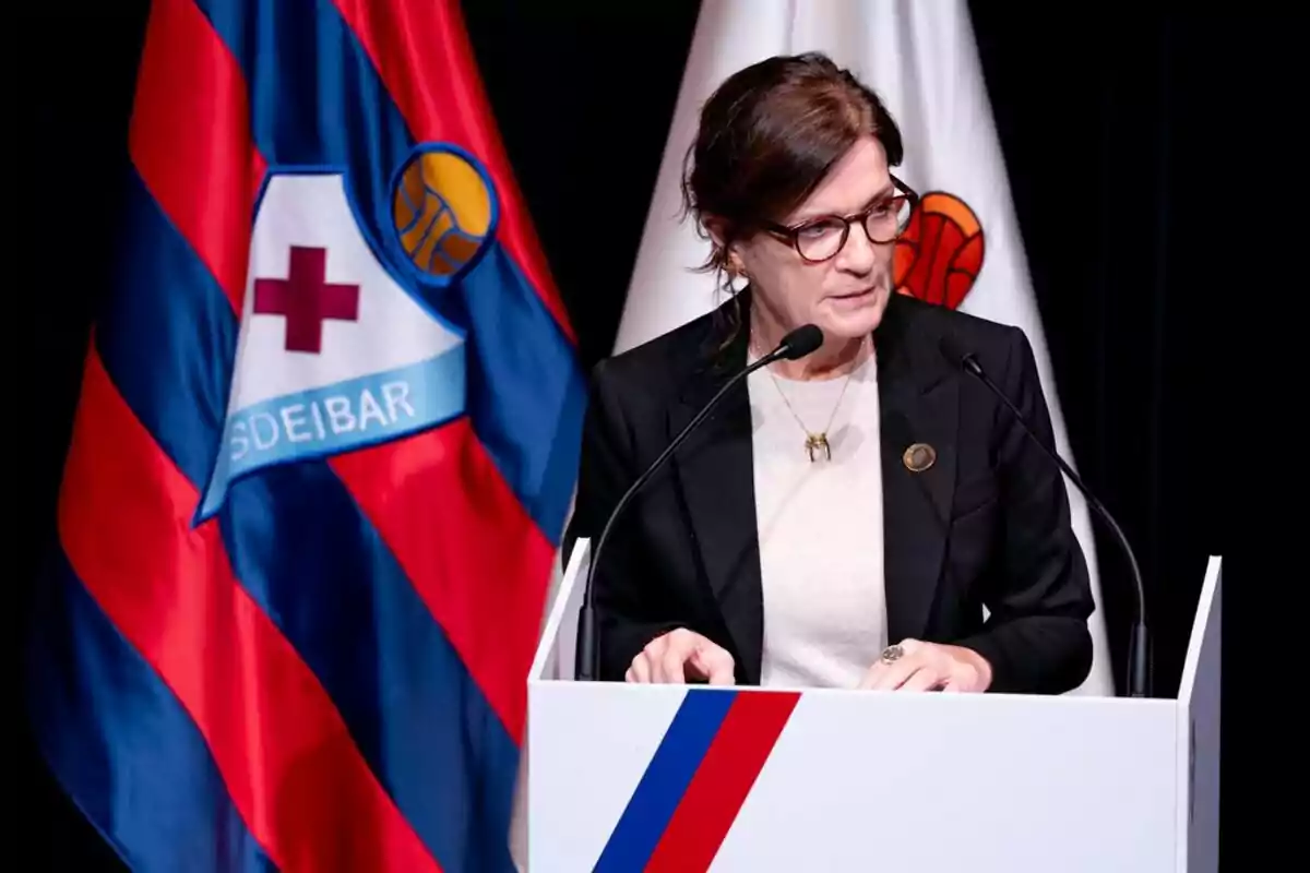 Una persona hablando en un podio con un micrófono y una bandera del SD Eibar al fondo. Una persona hablando en un podio con un micrófono y una bandera del SD Eibar al fondo.