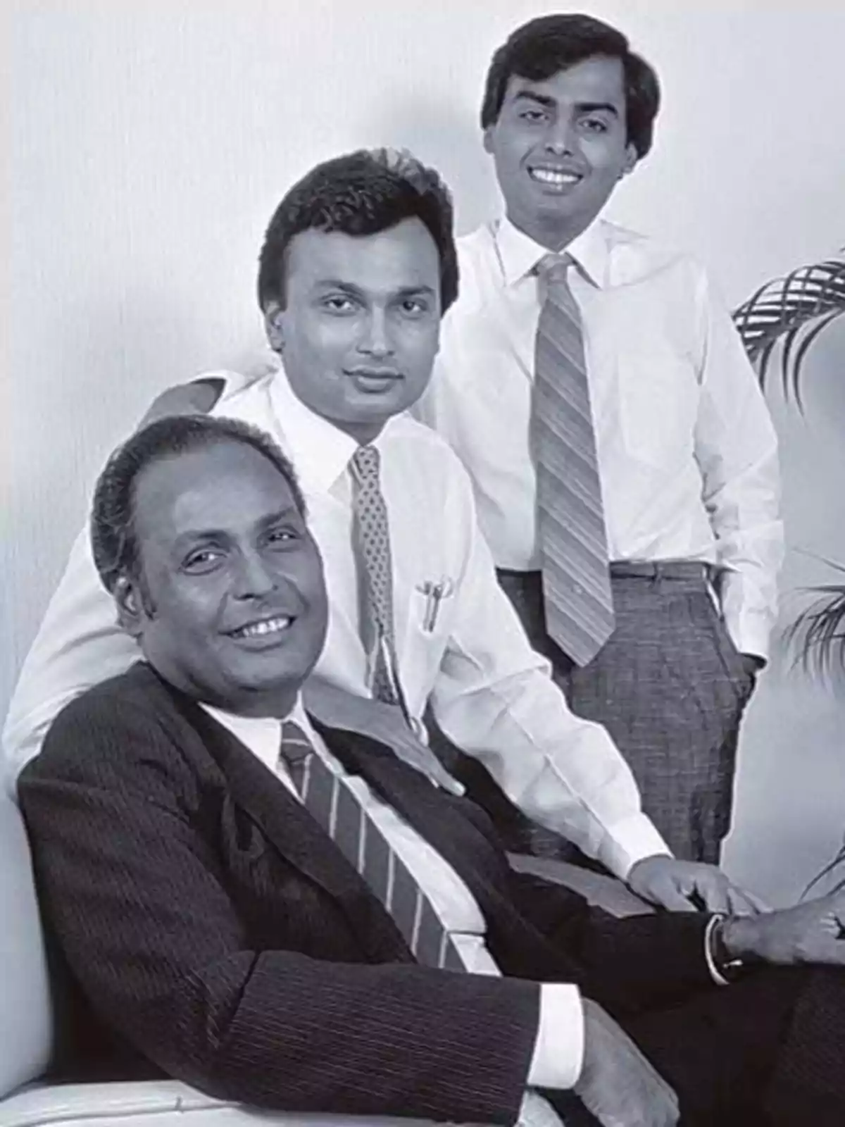 Tres hombres posando juntos, dos de pie y uno sentado, todos vestidos con camisas y corbatas.