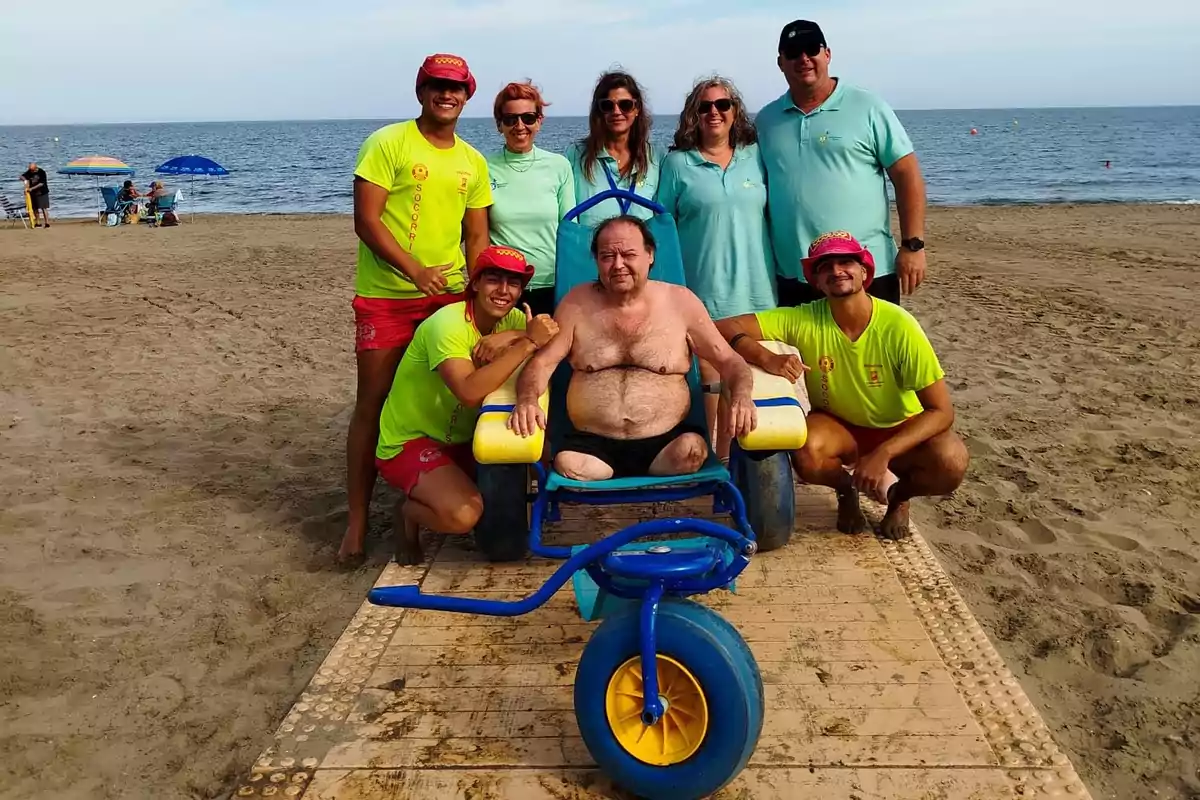 Un grupo de personas en la playa rodea a un hombre sentado en una silla de ruedas adaptada para la arena.