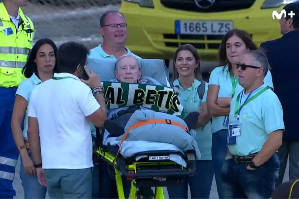 Un grupo de personas rodea a un hombre en una camilla que sostiene una bufanda del Real Betis mientras sonríen y conversan.