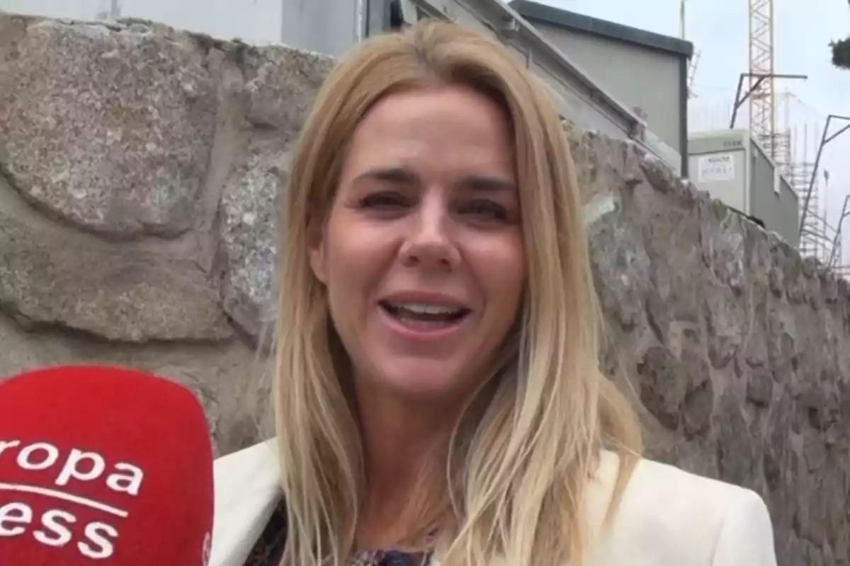 Amelia Bono sonríe frente a una pared de piedra mientras sostiene un micrófono rojo. Amelia Bono sonríe frente a una pared de piedra mientras sostiene un micrófono rojo.