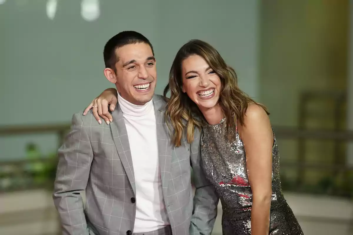 Dos personas sonrientes posan juntas, una con traje gris y la otra con vestido brillante.