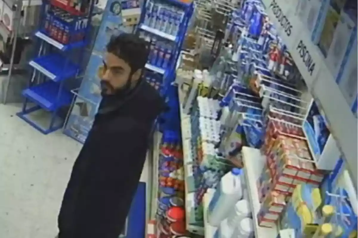 Un hombre con barba y cabello oscuro está de pie en una tienda, rodeado de estantes llenos de productos diversos, incluyendo artículos de limpieza y otros productos de consumo.