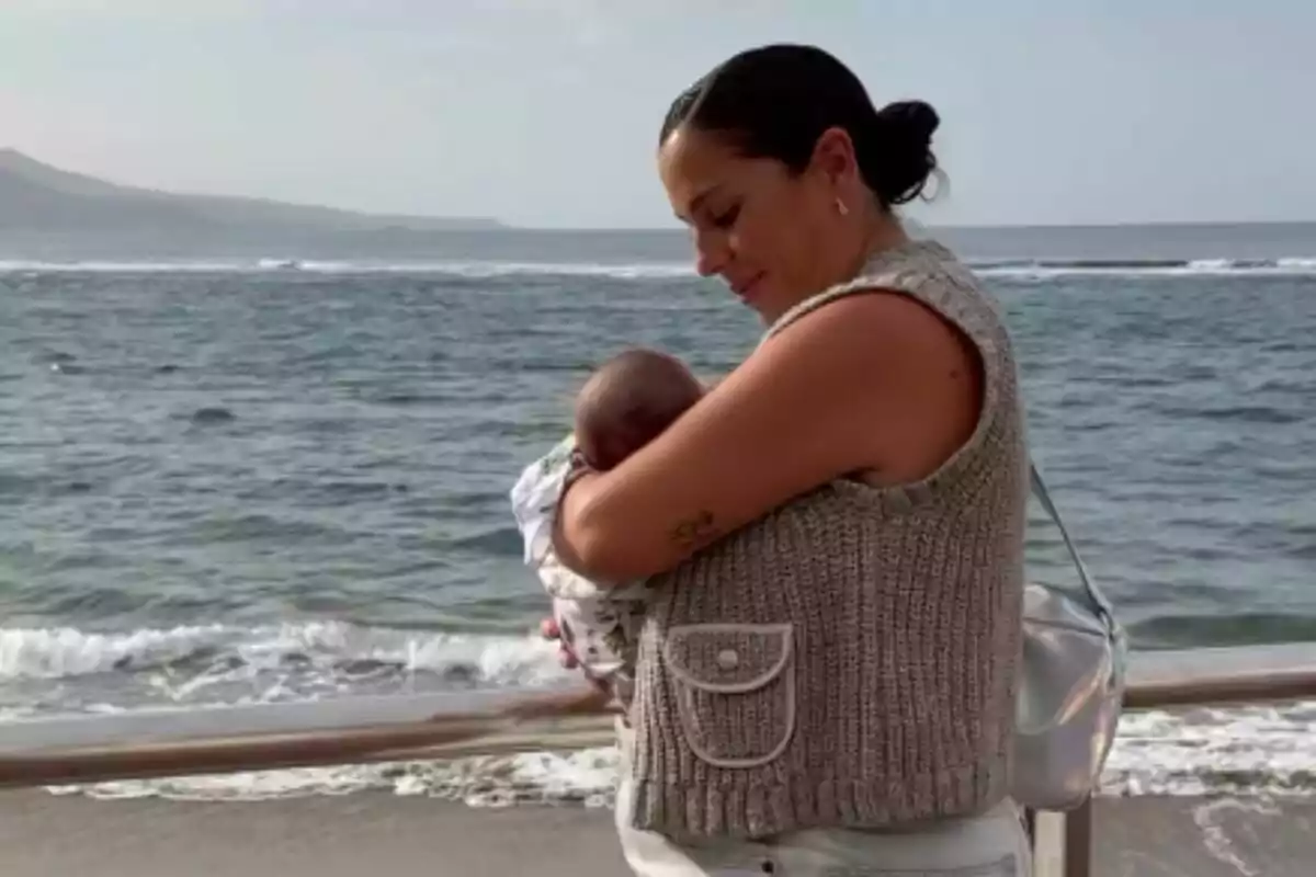 Plano medio de Anabel Pantoja mientras sostiene a un bebé -que es su hija Alma- mientras está de pie frente al mar. Plano medio de Anabel Pantoja mientras sostiene a un bebé -que es su hija Alma- mientras está de pie frente al mar.