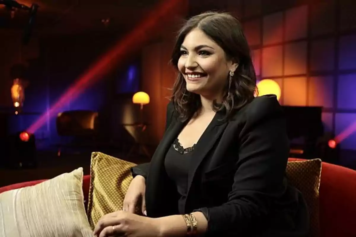 Una mujer sonriente sentada en un sofá con cojines en un ambiente iluminado con luces de colores.