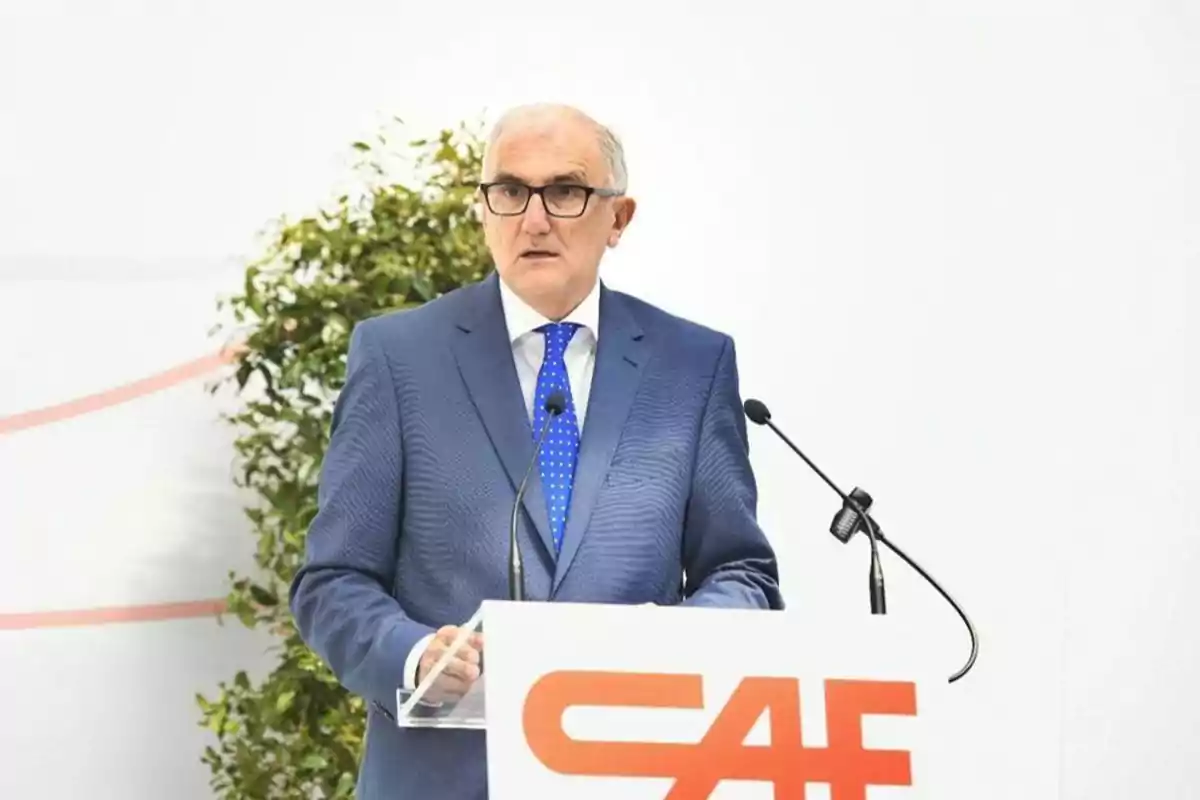 Un hombre con traje azul y corbata azul de lunares habla en un podio con el logo de CAF y un fondo blanco con plantas.