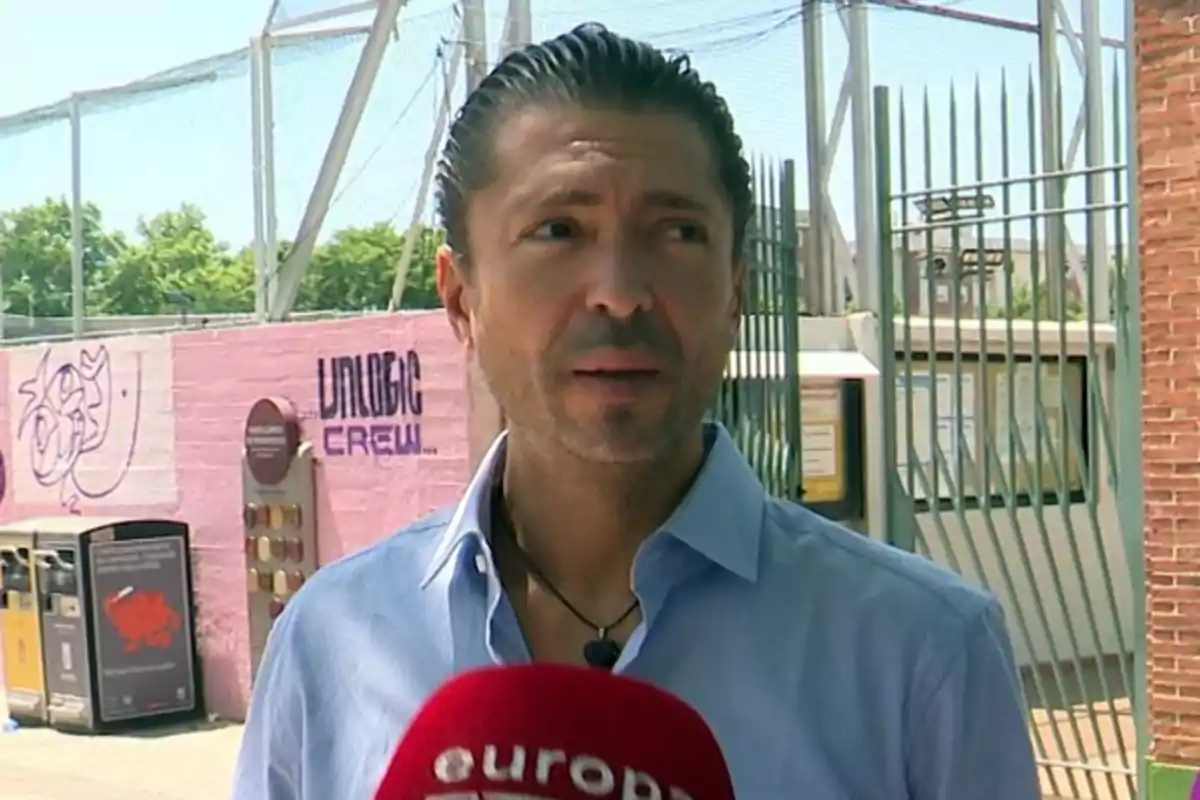 Un hombre con camisa azul es entrevistado frente a una reja y un muro con grafitis.