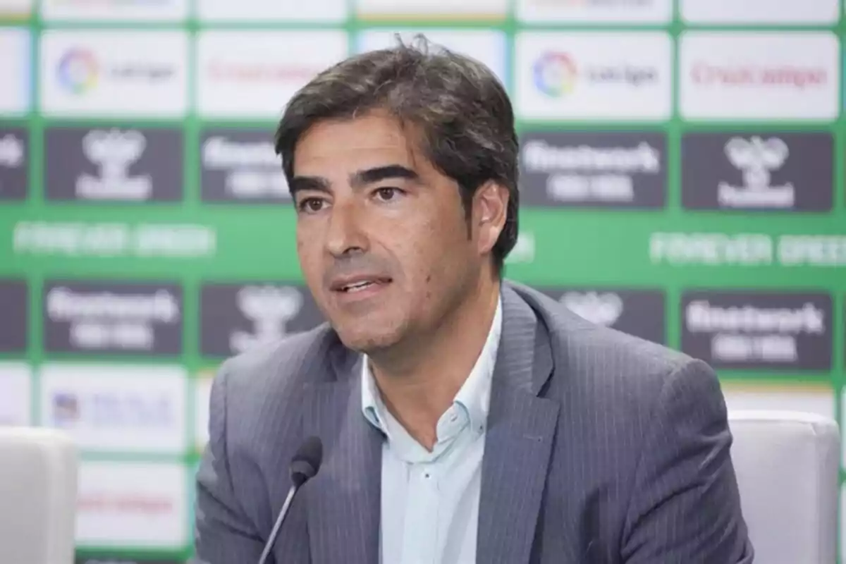 Un hombre en traje hablando en una conferencia de prensa con un fondo de logotipos deportivos. Un hombre en traje hablando en una conferencia de prensa con un fondo de logotipos deportivos.