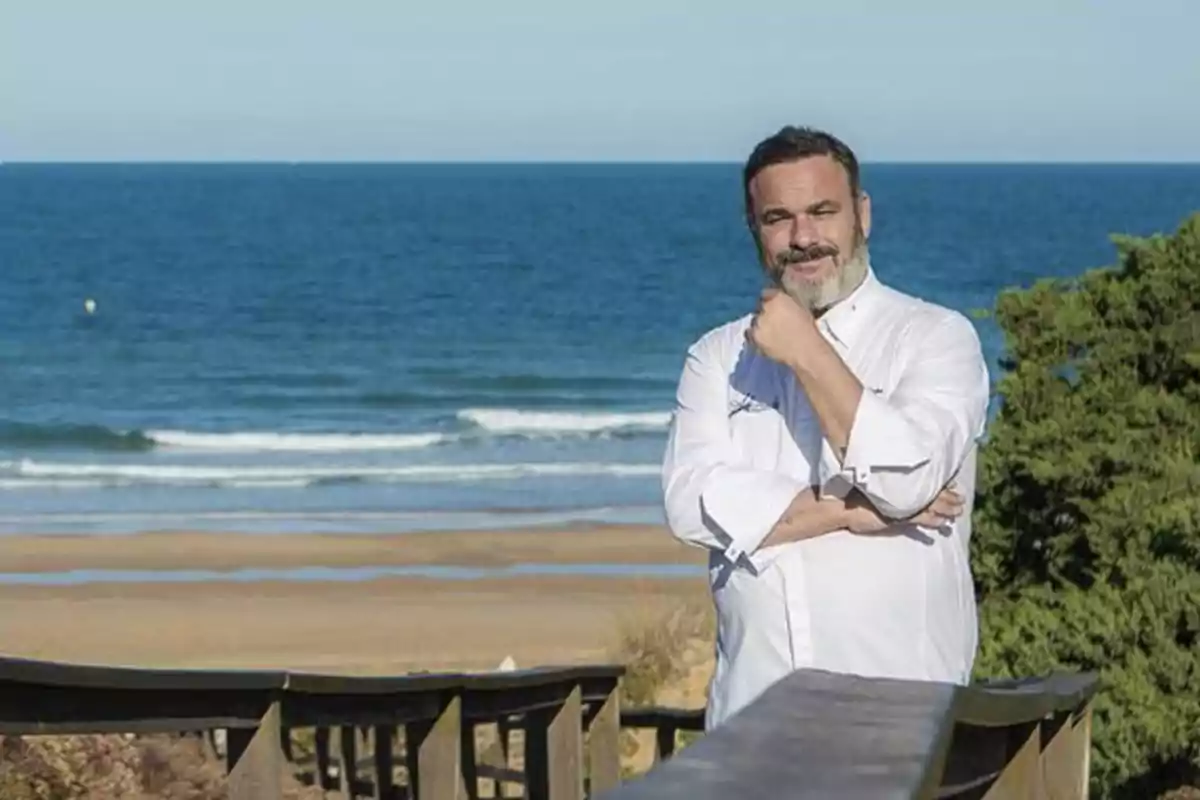 Un hombre con uniforme de chef posando en una pasarela de madera frente al mar.