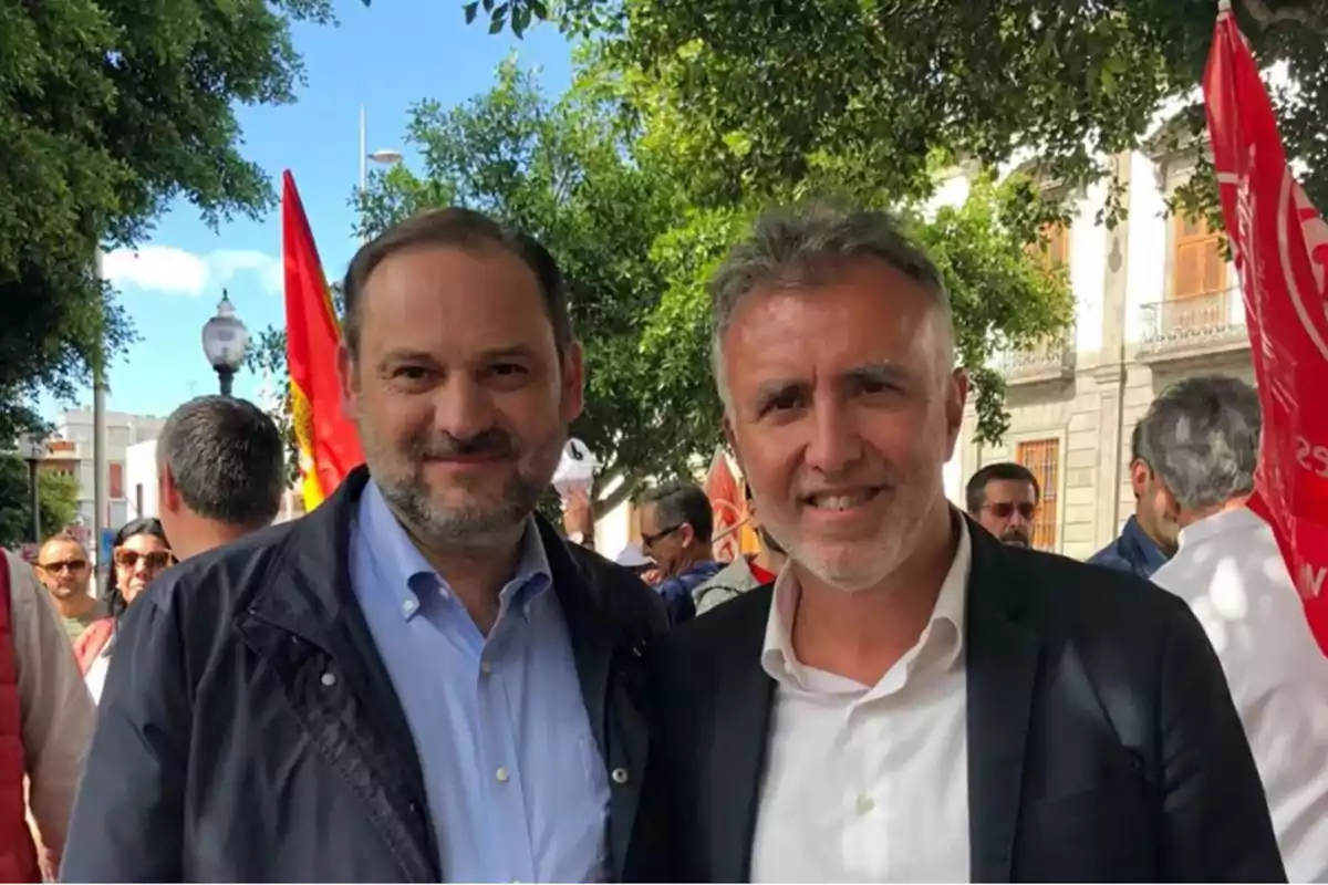 Dos hombres posan juntos en un evento al aire libre con banderas rojas y árboles en el fondo.