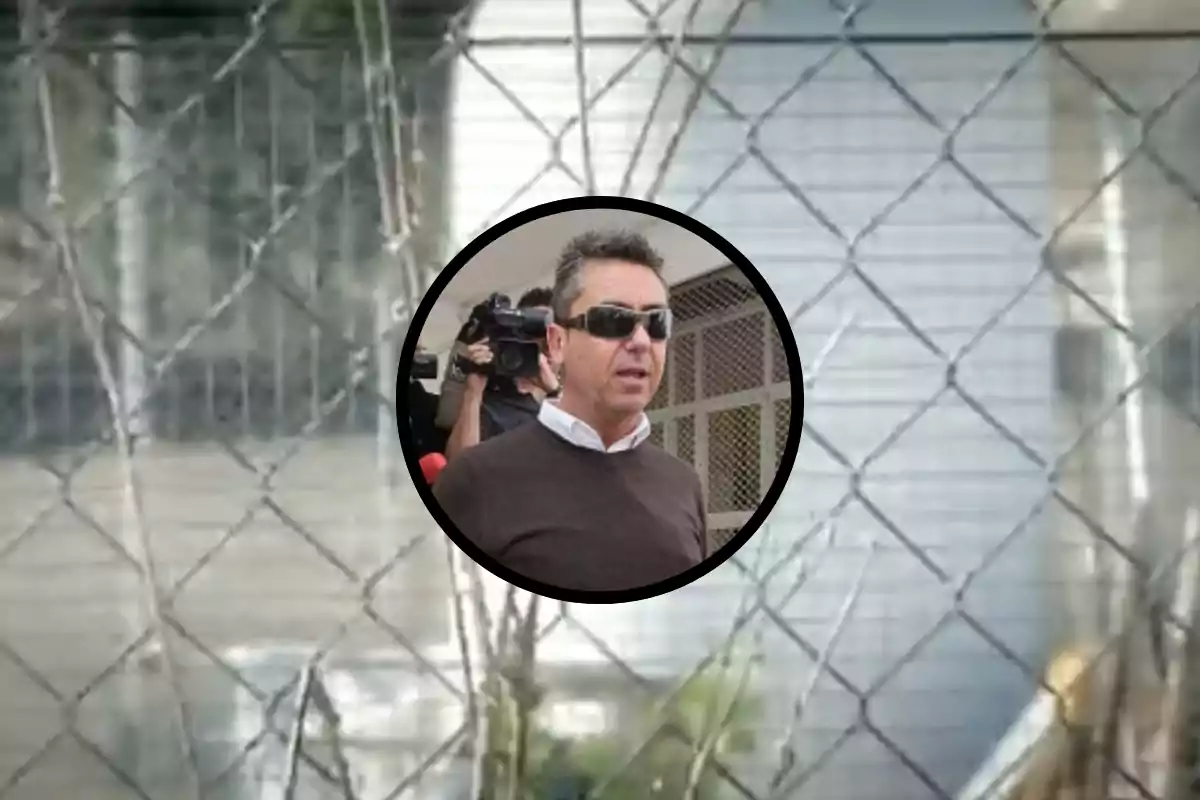 Un hombre con gafas de sol y un suéter oscuro está siendo fotografiado por un camarógrafo mientras se encuentra frente a un edificio con una cerca de alambre. Un hombre con gafas de sol y un suéter oscuro está siendo fotografiado por un camarógrafo mientras se encuentra frente a un edificio con una cerca de alambre.