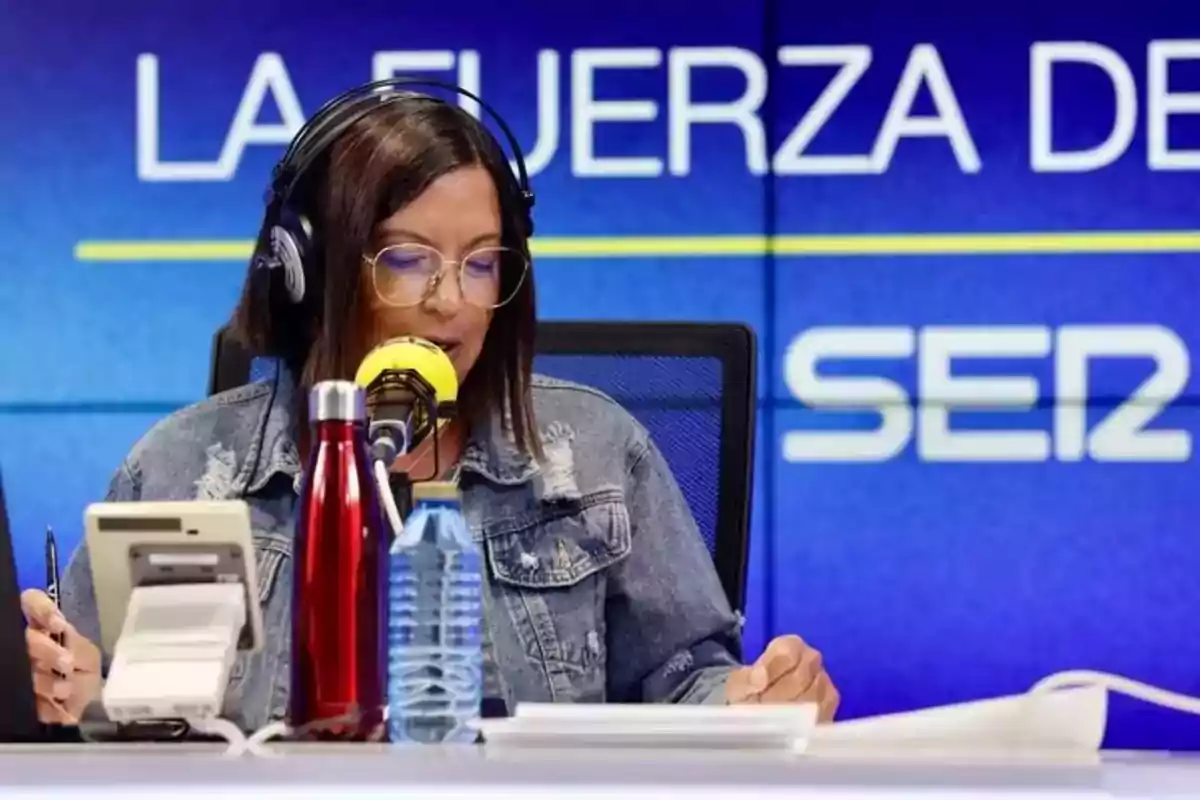 Una mujer con gafas y auriculares habla frente a un micrófono amarillo en un estudio de radio con el logo de la cadena SER en el fondo.