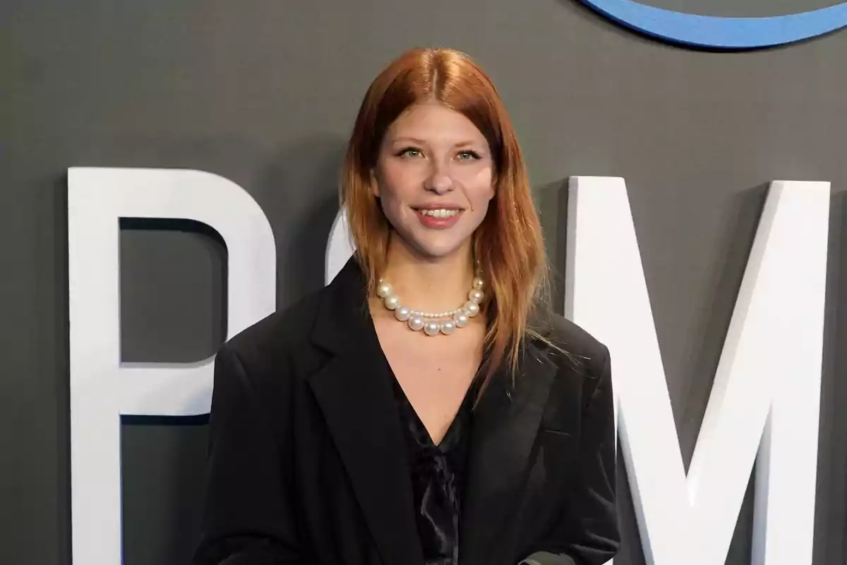 Una persona con cabello pelirrojo y collar de perlas, vestida con un traje negro, posa frente a un fondo con letras grandes.