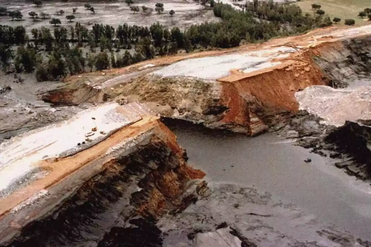 Vista aérea de un colapso de una presa de relaves mineros, con agua y lodo esparcidos por el área circundante y árboles en el fondo.