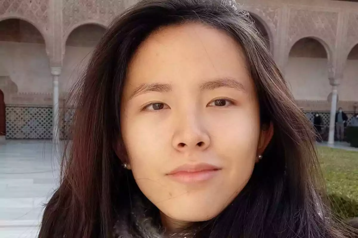 Una mujer con cabello largo y oscuro, posando frente a un edificio con arcos y detalles arquitectónicos intrincados.