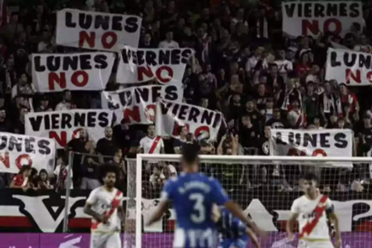 Un grupo de aficionados en un estadio sostiene pancartas que dicen "Lunes NO" y "Viernes NO" mientras se desarrolla un partido de fútbol.