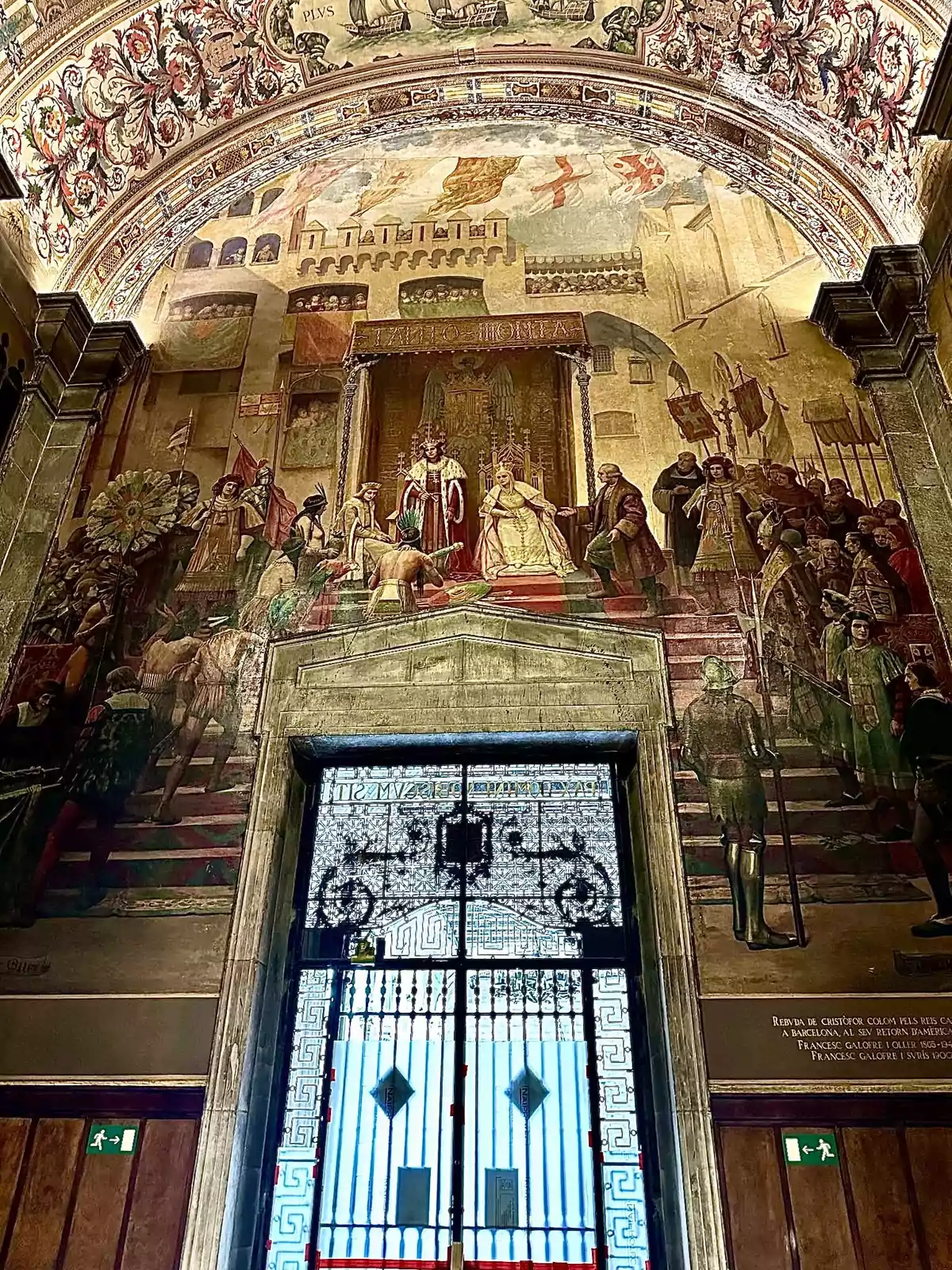 Una pintura mural detallada y colorida que representa una escena histórica con figuras reales y personajes en un entorno arquitectónico ornamentado, con una puerta de hierro forjado en la parte inferior.