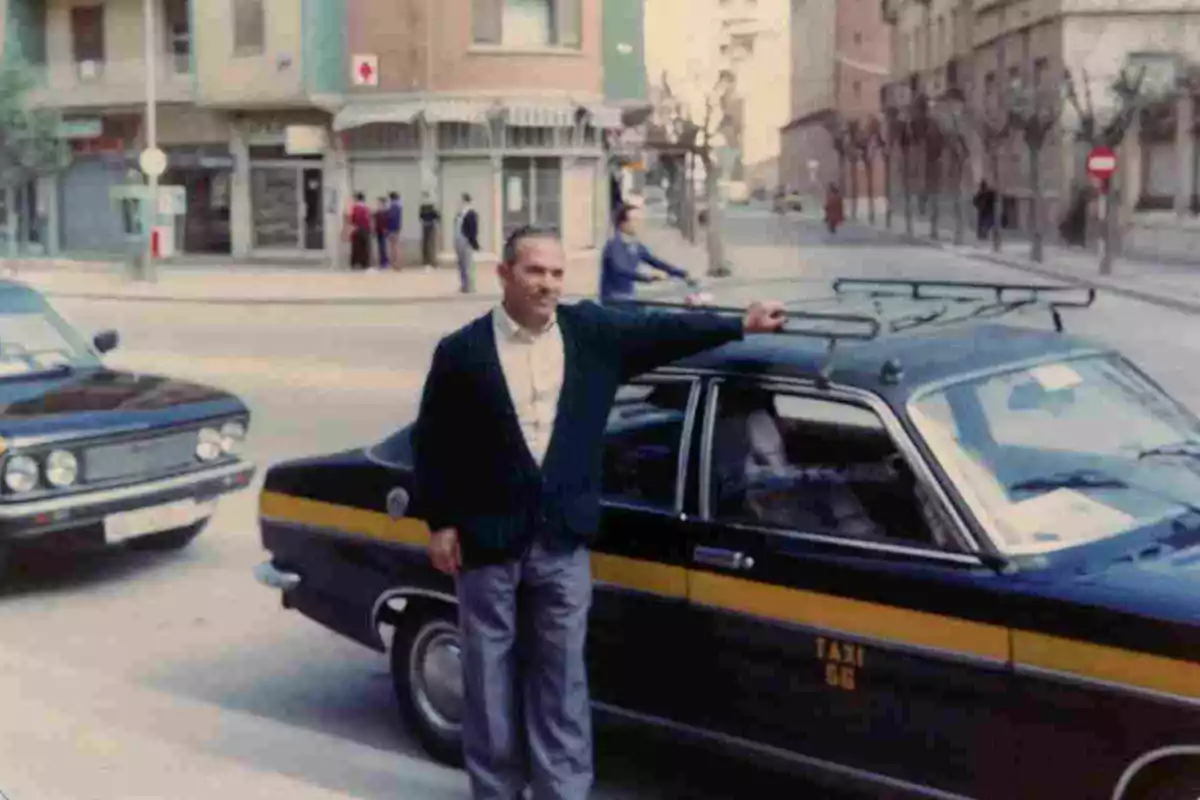 Un hombre posa junto a un taxi en una calle urbana con edificios y personas al fondo.