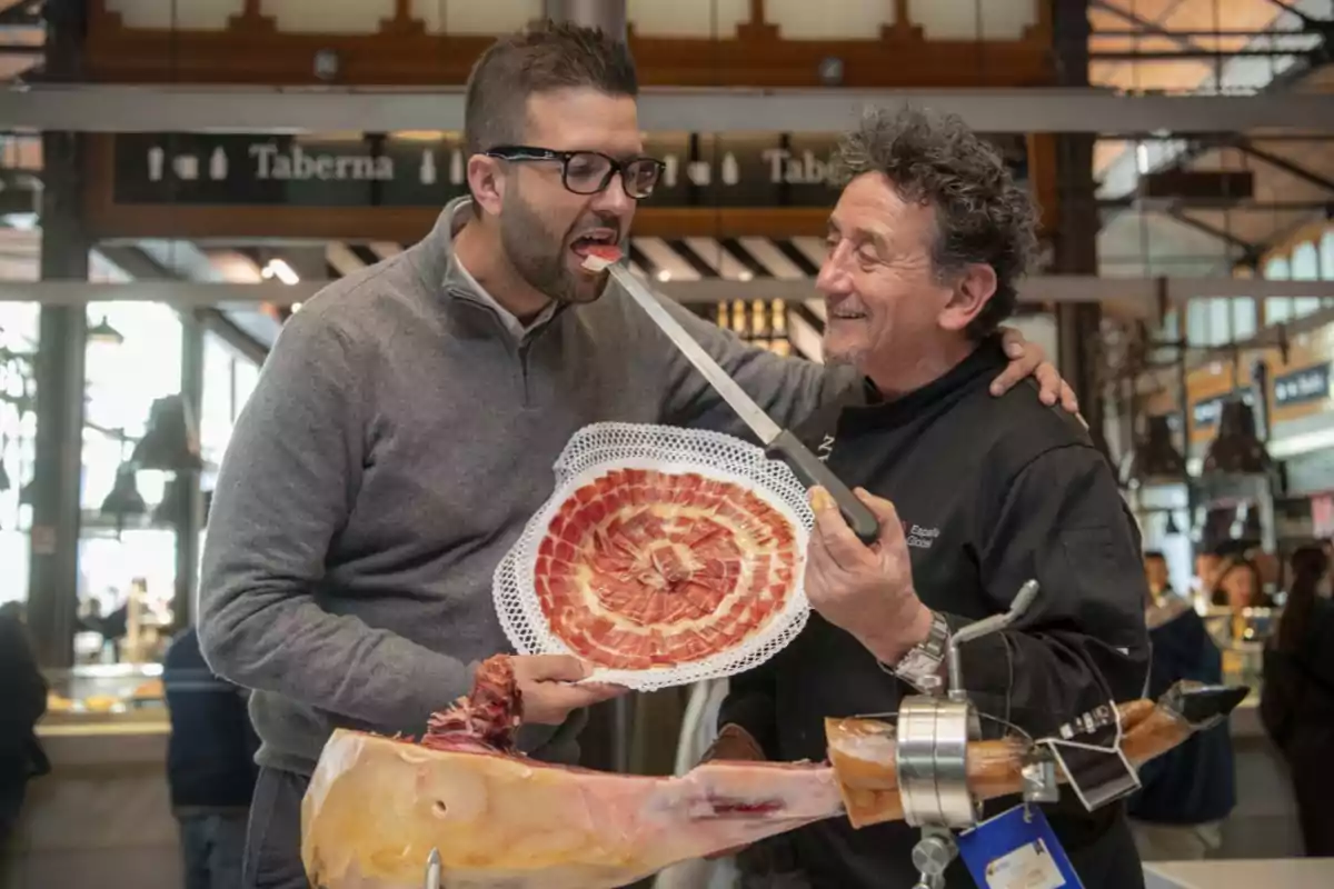 Dos hombres sonrientes en un mercado, uno de ellos sostiene un plato de jamón y el otro le ofrece una rebanada con un cuchillo.