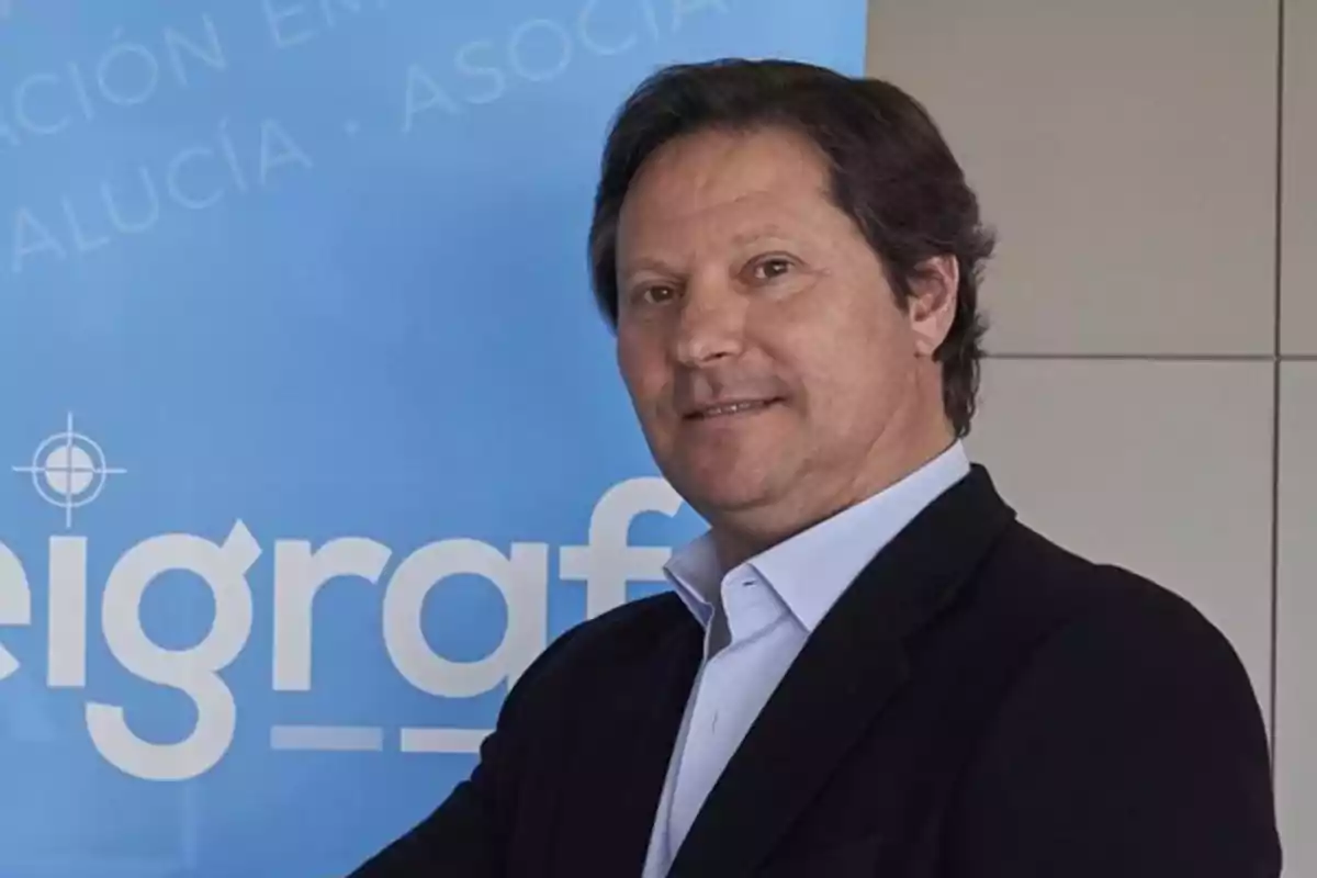 Hombre de cabello castaño y traje oscuro posando frente a un cartel azul con letras blancas Hombre de cabello castaño y traje oscuro posando frente a un cartel azul con letras blancas