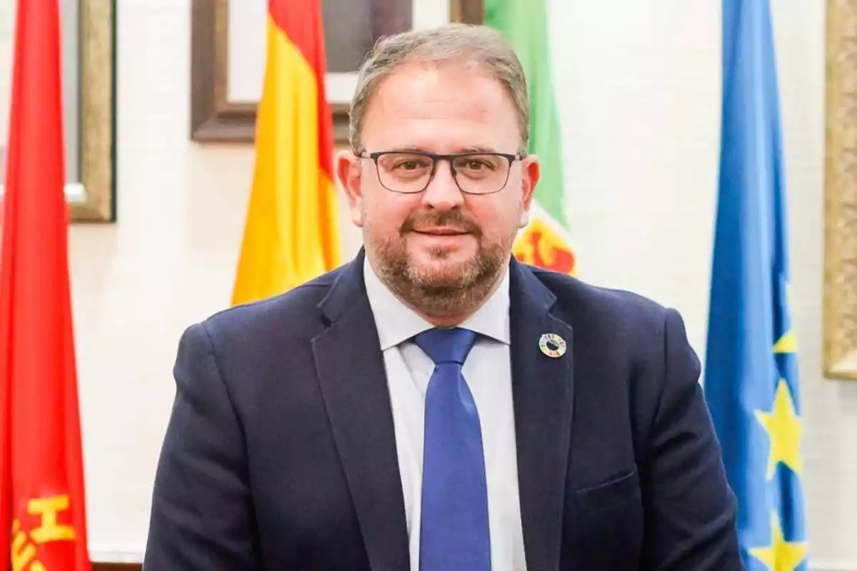 Hombre con gafas y traje oscuro posando frente a varias banderas.