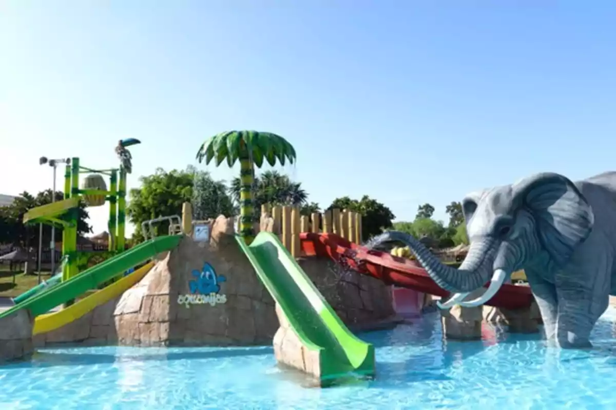 Parque acuático infantil con toboganes de colores, una palmera artificial y una figura de elefante junto a una piscina bajo un cielo despejado