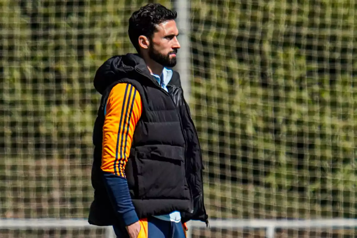 Un hombre con barba y cabello oscuro lleva una chaqueta acolchada negra sobre una sudadera deportiva naranja con rayas azules mientras está de pie frente a una red de fútbol. Un hombre con barba y cabello oscuro lleva una chaqueta acolchada negra sobre una sudadera deportiva naranja con rayas azules mientras está de pie frente a una red de fútbol.