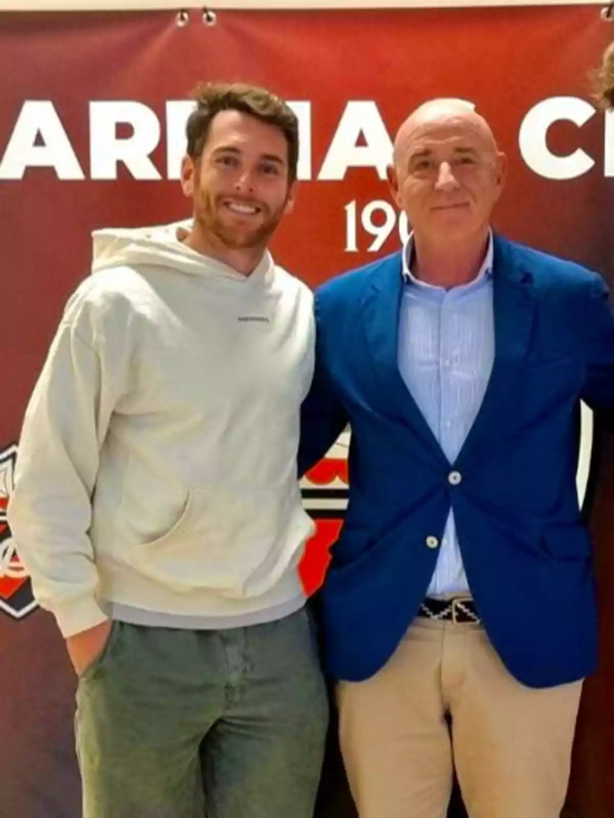 Dos personas posan juntas frente a un cartel rojo.