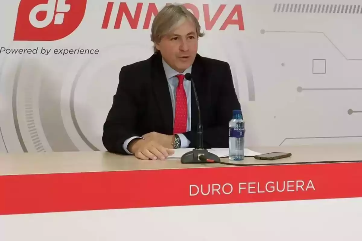 Hombre en traje hablando en una conferencia de prensa de Duro Felguera con una botella de agua y un teléfono sobre la mesa.